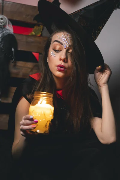 魔女の衣装の女の子は片手で彼女の帽子を保持し 一方でろうそくが燃える頭蓋骨の形をしたキャンドルホルダー 魔女は火に吹く ハロウィーンの雰囲気 — ストック写真