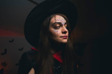 Siyah cadı şapkalı genç bir kız. Yüzünde pullar var. Karnaval kostümü ve Cadılar Bayramı partisiyle ilgileniyor.