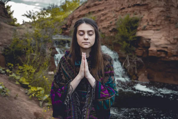 young beautiful girl with closed eyes in an Indian poncho who meditates outdoors near a small waterfall, reuniting with nature