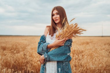 Kot ceketli ve beyaz elbiseli güzel bir kız, tatlı tatlı gülümsüyor, olgun çavdar kulaklarını elinde tutuyor, sonbahar buğday tarlasında duruyor.,