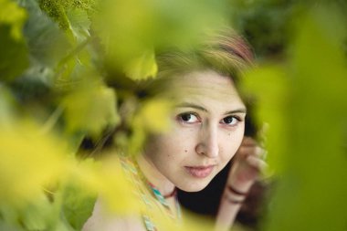 Genç kız yeşil asma yapraklarından kameraya bakıyor.