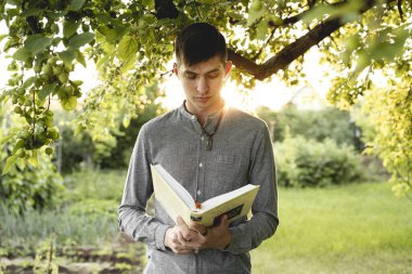 Elinde kitap olan genç bir adam yeşil bir bahçede duruyor.