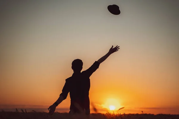 silhouette of a man standing in a field who catches his flying hat with his hand outstretched at sunset, blue sky, freedom, close-up