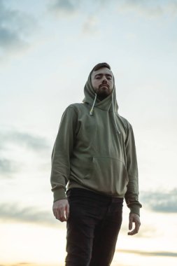 Yeşil tişörtlü genç adam günbatımı gökyüzünün arka planında duruyor, gri sirrus bulutları, doğayla birleşiyor, dikey yönelim.