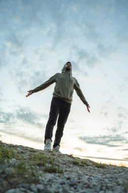 Yeşil eşofmanlı genç bir adam kollarını farklı yönlere açmış günbatımı gökyüzünün arka planına doğru uzanıyor. Gri sirus bulutları doğayla yeniden birleşiyor.