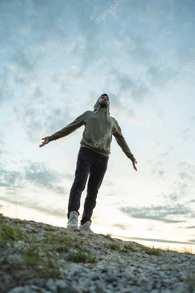 A young man in a green sweatshirt stands with his arms outstretched in different directions against the background of the sunset sky, gray cirrus clouds, reuniting with nature
