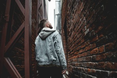 Kot ceketli genç sakallı adam tuğla bir sokakta yürür ve yukarı bakar, yeraltına.