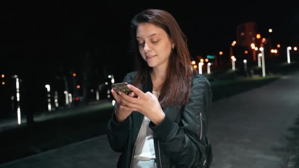 Uma bela mulher sociável fica em um parque noturno, mensagens de texto e flertando — Vídeo de Stock