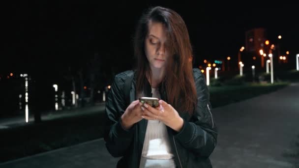 Uma bela mulher sociável fica em um parque noturno mensagens de texto por telefone — Vídeo de Stock