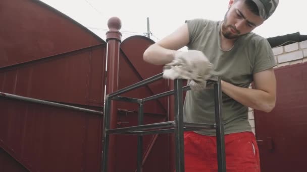 Een jonge man in een werkend uniform en handschoenen veegt een metalen frame grondig af — Stockvideo