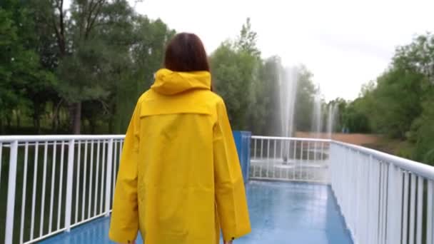 Chica en un impermeable amarillo camina a través del puente y se pone una capucha. — Vídeo de stock