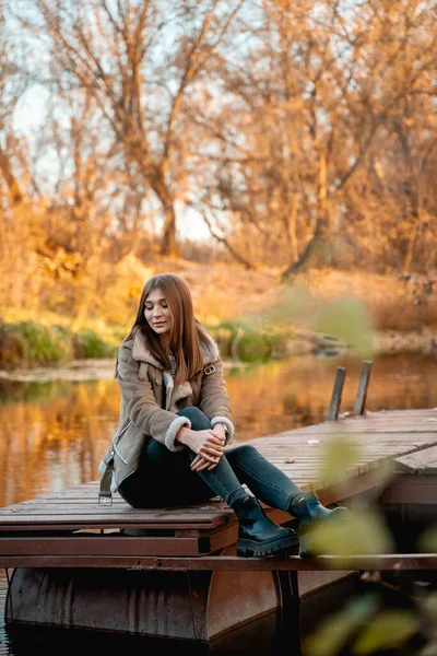 Όμορφη γυναίκα κάθεται δίπλα στη λίμνη και απολαμβάνει την ομορφιά του χρυσού φθινοπώρου — Φωτογραφία Αρχείου