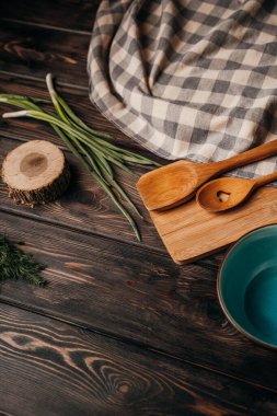 Mutfak gereçleri ahşap arka planda yatar, üst manzara, kesme tahtası, kaşık, spatula, tabak, maydanoz ve yeşil soğan.