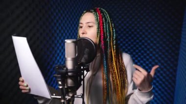 Renkli örgülü kız kayıt stüdyosunda mikrofona rap yapıyor.