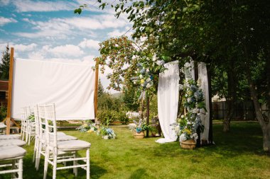Açık havada güzel bir düğün töreni. Madalyalı sandalyeler çimlerin üzerinde duruyor. Yeşil doğal arka planda kumaş ve beyaz çiçeklerden yapılmış düğün kemeri.