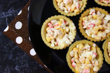 Yengeç etli tart, konserve mısır ve yumurta, siyah tabakta ve peçetede, merkezin sağında, en üstte.