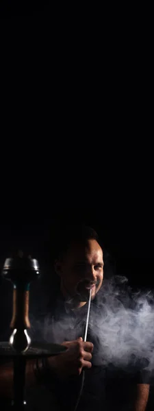 男と縦のバナー伝統的なフックパイプ喫煙 コピースペースと黒の背景に煙を吐き出す男 フックバーの喫煙コンセプト テキストの場所 — ストック写真