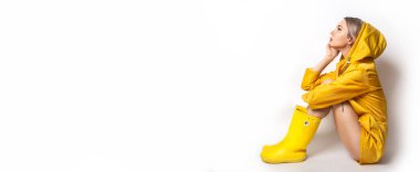 Sarı yağmurluk ve lastik çizme giyen genç bir bayrağın olduğu yatay pankart. Beyaz arka planda oturan moda sarışın kız. Bahar ve sonbahar giysileri