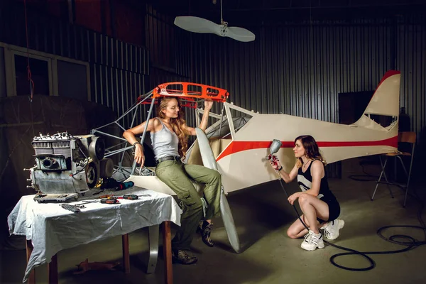 Two young women painting a body plane in airplane hangar.
