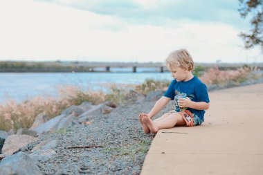 Küçük çocuk nehir kenarında oturuyor. Arka planda köprü var.