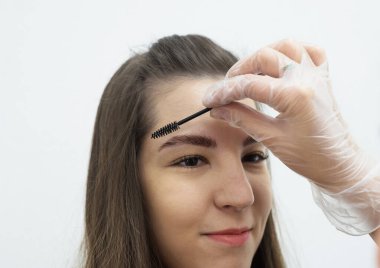 Stilist güzellik salonunda güzel bir kadın için profesyonel makyaj yapıyor. Eldiven ustası hastaların kaşlarını özel bir fırçayla tarıyor. 