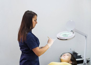 Kalıcı kaş makyajı sırasında salonda usta ve müşteri var. Elinde kalem ve klasör olan bir usta bilgi yazar..