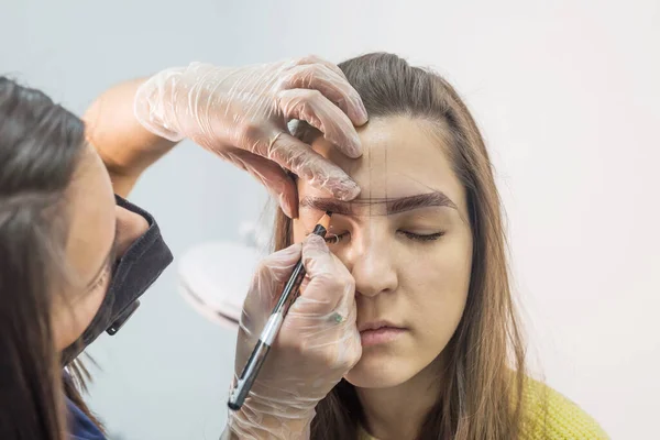 eyebrow master draws the shape of the eyebrows of a young woman with an eyebrow pencil. Henna eyebrow coloring procedure in a beauty salon