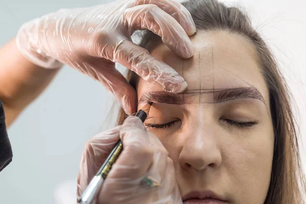 eyebrow master draws the shape of the eyebrows of a young woman with an eyebrow pencil. Henna eyebrow coloring procedure in a beauty salon