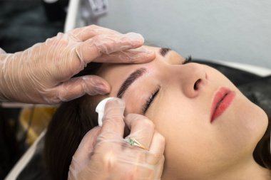 Güzellik salonunda, uzman bir kozmetik uzmanı Avrupalı bir kız için kalıcı makyaj yapar, kaşlarını pamukla siler. İşlem sırasında modelin yüzünün yakın çekimi.