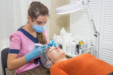 Kız kozmetik uzmanı, kozmetoloji ofisindeki hastasına ultrasonik yüz temizleme prosedürünü uyguluyor.