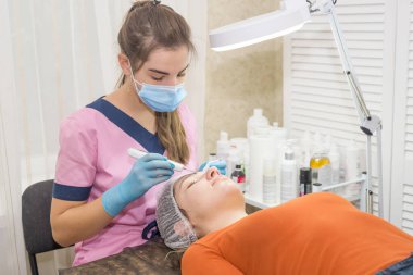 Kız kozmetik uzmanı, kozmetoloji ofisindeki hastasına ultrasonik yüz temizleme prosedürünü uyguluyor.