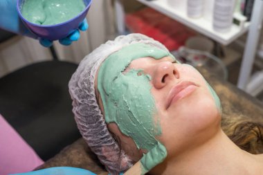 Esmer bir kız spa salonunda işlem görüyor, mavi eldivenli usta bir güzellik uzmanı yeşil bir maskenin kalın bir katmanını uyguluyor, derisini pislikten temizliyor, yüzüne göz kulak oluyor..