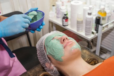 Esmer bir kız spa salonunda işlem görüyor, mavi eldivenli usta bir güzellik uzmanı yeşil bir maskenin kalın bir katmanını uyguluyor, derisini pislikten temizliyor, yüzüne göz kulak oluyor..