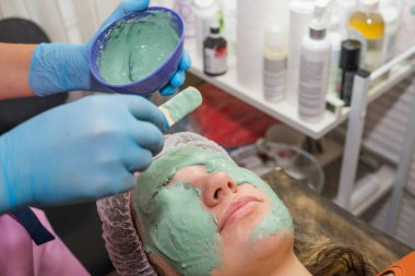 Esmer bir kız spa salonunda işlem görüyor, mavi eldivenli usta bir güzellik uzmanı yeşil bir maskenin kalın bir katmanını uyguluyor, derisini pislikten temizliyor, yüzüne göz kulak oluyor..