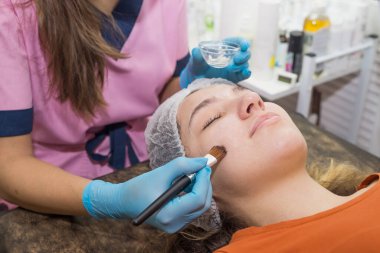 Güzellik uzmanı yüz maskesi takıyor. Genç bir kız kanepede yatıyor. Güzellik salonunda kozmetik prosedürleri.