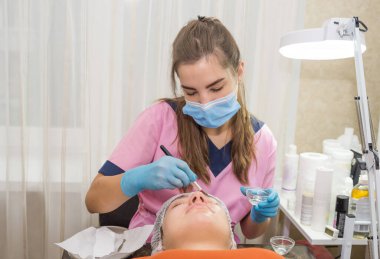 Olumlu genç bir kadın gözleri kapalı yatıyor. Güzellik uzmanı beyaz kremalı nemlendirici bir maske takıyor. Güzellik ve cilt bakımı