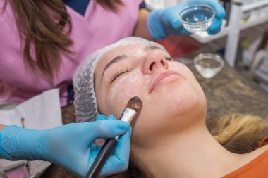 Olumlu genç bir kadın gözleri kapalı yatıyor. Güzellik uzmanı beyaz kremalı nemlendirici bir maske takıyor. Güzellik ve cilt bakımı