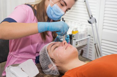Olumlu genç bir kadın gözleri kapalı yatıyor. Güzellik uzmanı beyaz kremalı nemlendirici bir maske takıyor. Güzellik ve cilt bakımı