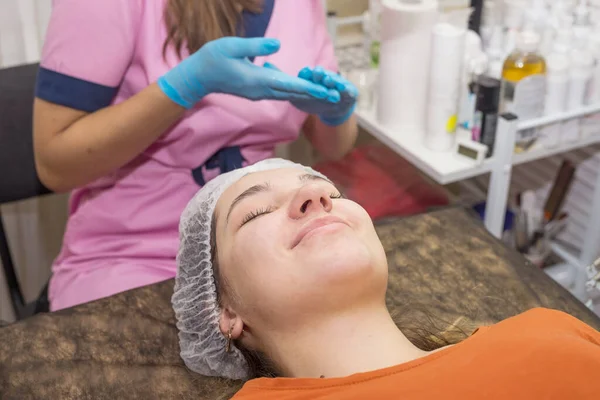 Beautician with gloves washes the face of a young woman. Removal of cosmetics. Cosmetic procedures in the beauty salon.