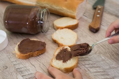 Elleri bir baget ekmeğe çikolata ezmesi sürüyor. Ahşap arkaplan.
