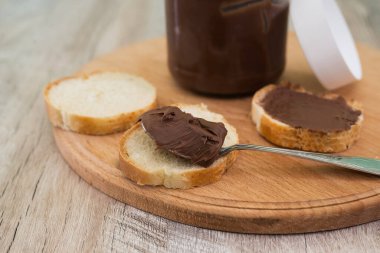 Bir kavanoz çikolatalı fındık ezmesi ve tahta arka planda baget dilimleri..