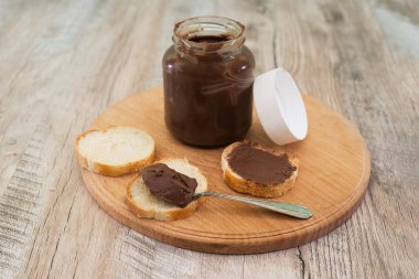 Bir kavanoz çikolatalı fındık ezmesi ve tahta arka planda baget dilimleri..