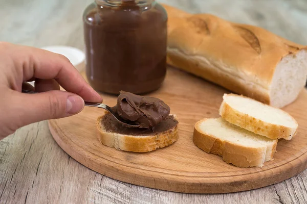 Elleri bir baget ekmeğe çikolata ezmesi sürüyor. Ahşap arkaplan.