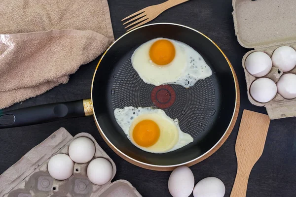 Fried eggs cooking process. Two fried eggs in a pan and raw eggs on a dark wooden background.
