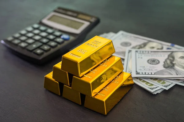 gold bars with dollars and a calculator on a black wooden background.