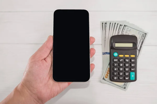 Female hand with a smartphone, dollars, calculator on a white wooden background. Copy space.