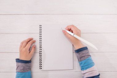 Masaüstü. Eller beyaz ahşap bir arka plana boş bir defter yazıyorlar. Düz konum, üst görünüm, tebrik kartı modeli, el işi not defteri ve kalem..