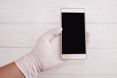 Beyaz eldivenli bir doktorun elinde beyaz arka planda siyah ekranlı bir akıllı telefon var. Uzayı kopyala Metnin için yer.