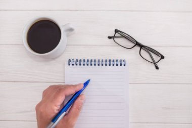 Kadın el beyaz ahşap arka planda boş bir deftere yazı yazıyor. Bardaklar ve bir fincan kahve. Düz yatıyordu. Yukarıdan görüntüle.