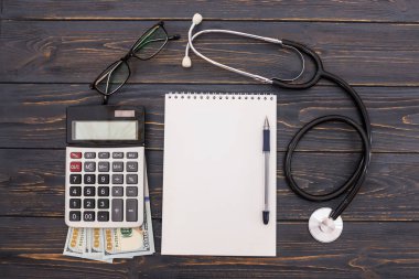 Boş not defteri, kalem, steteskop, hesap makinesi, ahşap arka planda dolar. Düz yatıyordu. Yukarıdan görüntüle.